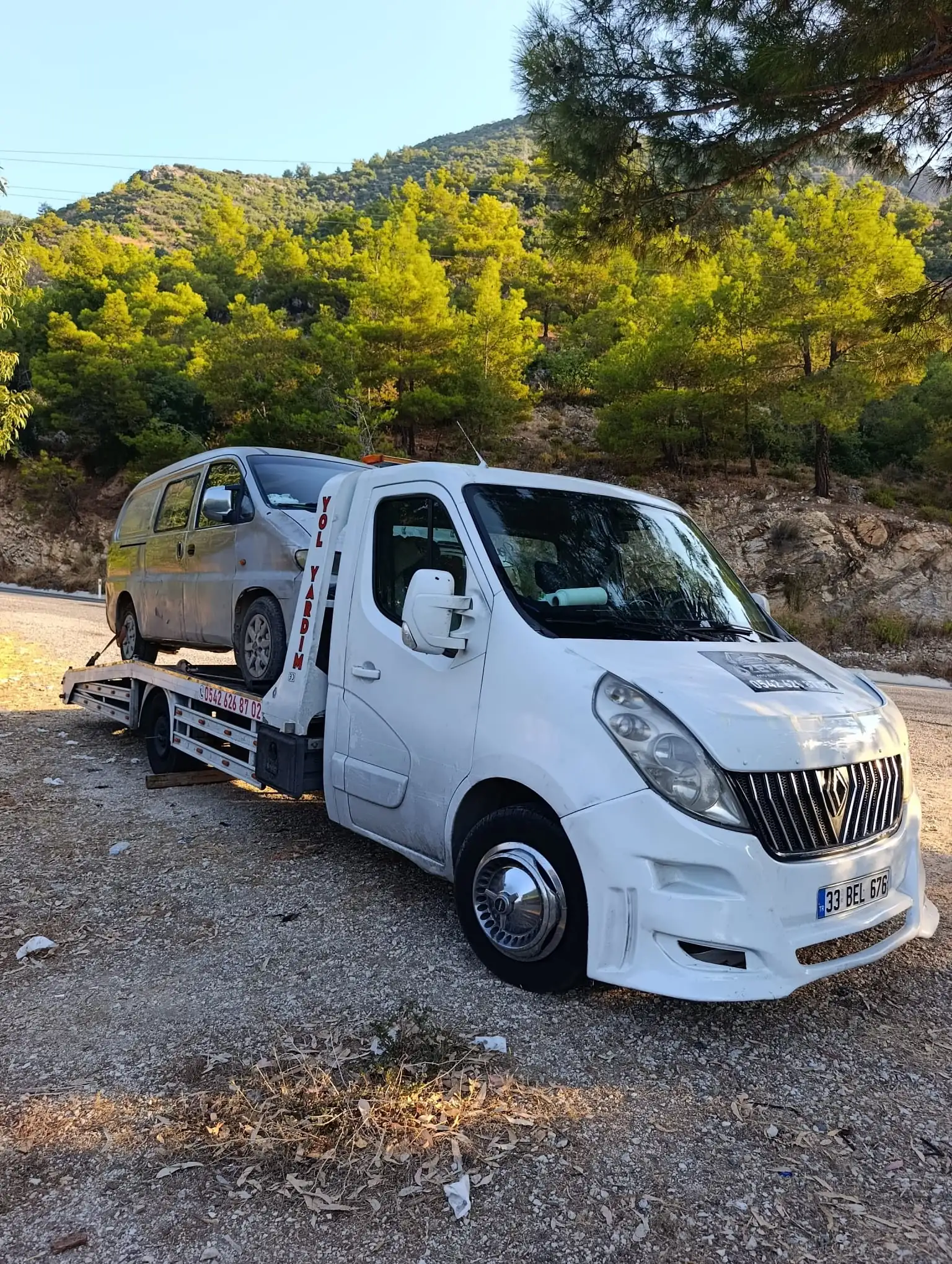 Ormanlık bir dağ yolunun kenarında, beyaz bir çekici gümüş rengi bir minibüsü kayar platformuna almış
