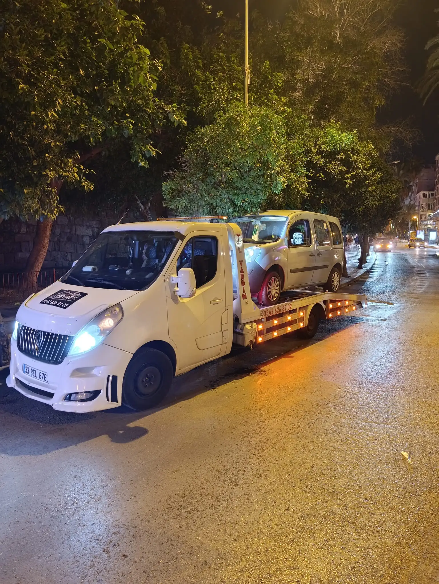 Gece bir caddenin kenarında, beyaz bir çekici gümüş rengi bir hafif ticari aracı kayar platformuna almış